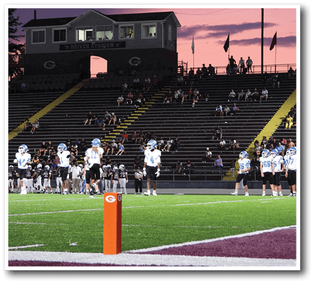 People in football uniforms stand on a green field near an orange marker with a stadium and spectators in the background under a purple sunset sky. Description generated by AI