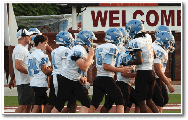 A group of people wearing blue helmets and white football uniforms are gathered on a field near a scoreboard and a sign that says welcome. Description generated by AI