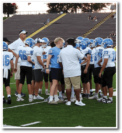 A group of people wearing blue and white football uniforms and helmets are gathered on a green field with empty bleachers in the background. Description generated by AI