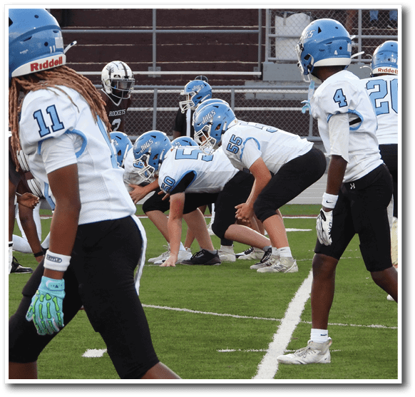 Five people wearing blue and white football uniforms are positioned on a football field preparing for a play while one person in a dark uniform stands opposite them. Description generated by AI