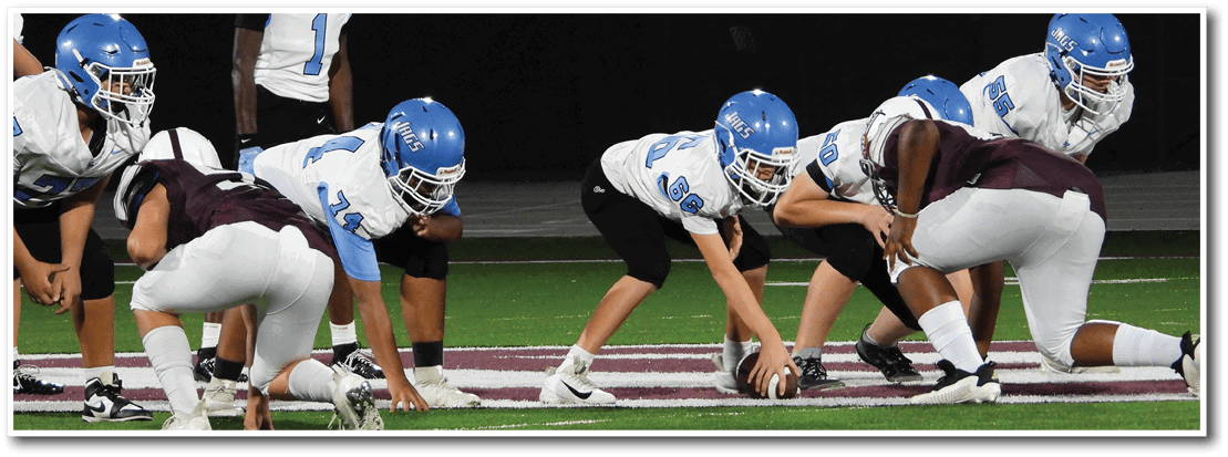 People in football uniforms are lined up on a field preparing for a play with one person holding the ball ready to snap it. Description generated by AI