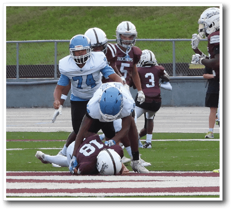A group of people wearing football uniforms are engaged in a play on a football field with one person lying on the ground and others standing or crouching nearby. Description generated by AI