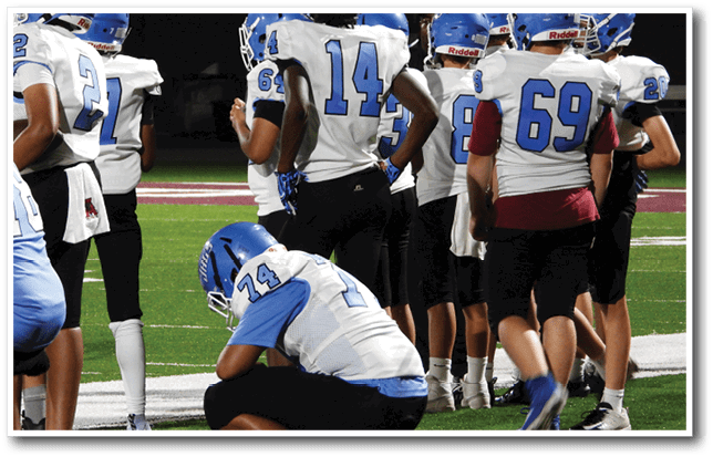 A person wearing a blue helmet and white sports jersey with the number 74 is crouched down on a football field while other people stand nearby. Description generated by AI