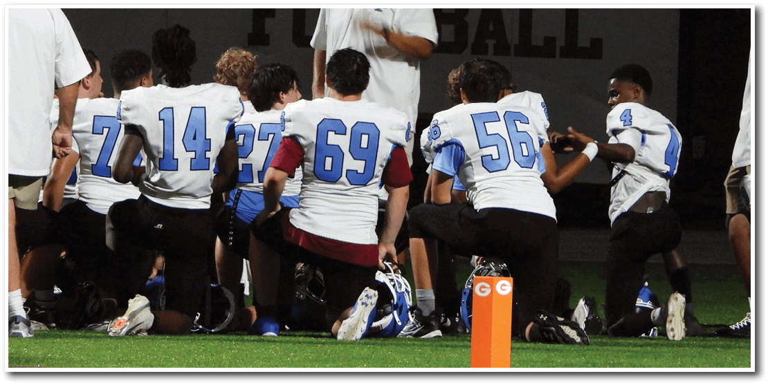 Several people wearing football uniforms with numbers kneel on a field near an orange marker during a nighttime event. Description generated by AI