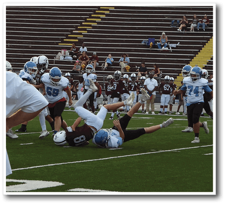 Two people in football uniforms are on the ground during a game, with one person rolling over and the other lying on their back on a green field. Description generated by AI