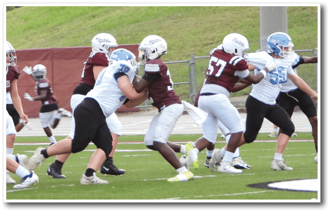 People wearing helmets and football uniforms are engaged in a play on a grassy football field with a fence and grassy hill in the background. Description generated by AI
