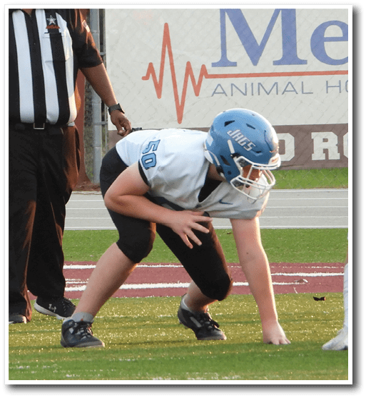 A person in a referee uniform stands on a football field near a player in a blue helmet and white jersey who is crouched in a ready position. Description generated by AI