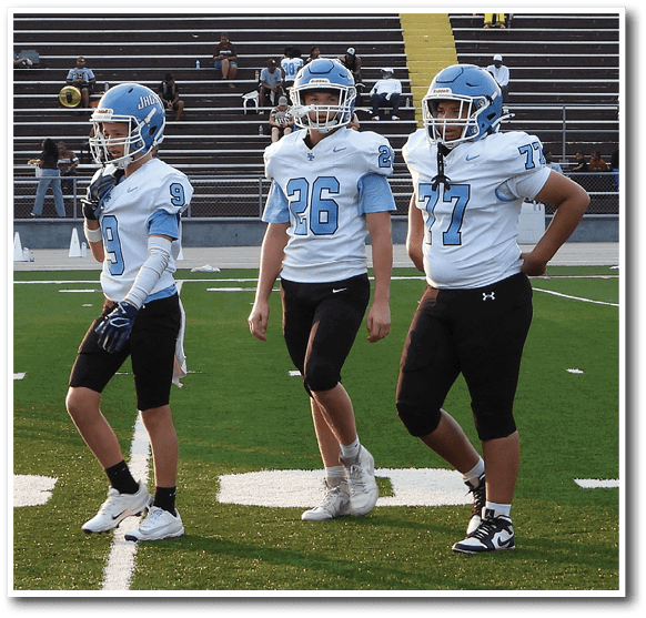 Three people in football uniforms and helmets stand on a field with bleachers and spectators in the background. Description generated by AI