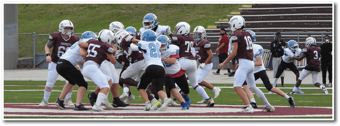 People wearing helmets and sports uniforms are engaged in a football game on a field with bleachers in the background. Description generated by AI