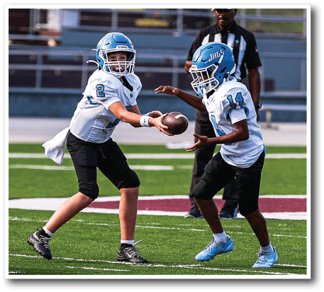 Two children wearing football helmets and uniforms are on a field with one handing the football to the other while a referee stands in the background. Description generated by AI