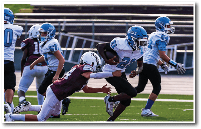 A group of people wearing football helmets and uniforms are playing on a field with one person running while holding the ball and another person reaching to tackle them. Description generated by AI