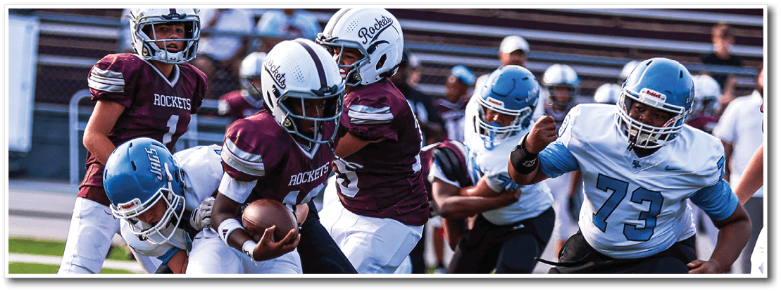 People in football uniforms are actively engaged in a play on a football field with one person holding the ball while others attempt to tackle or block. Description generated by AI