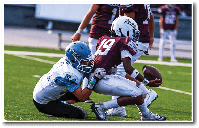 People wearing football uniforms are engaged in a play on a football field with one player holding the ball while others attempt to tackle. Description generated by AI