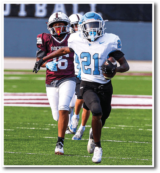 Two people in football uniforms are running on a field with one person holding a football and the other chasing behind. Description generated by AI