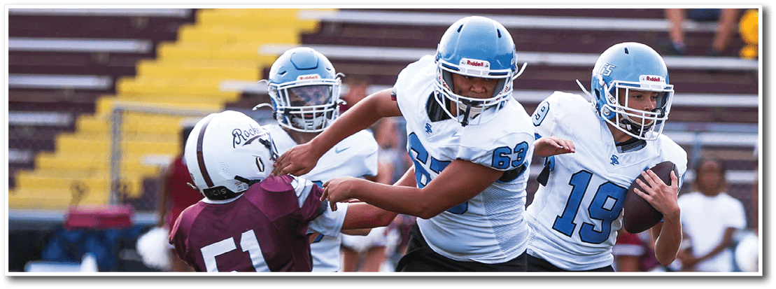 People in football uniforms are engaged in a play on a field with one person holding the ball and others blocking or attempting to tackle. Description generated by AI