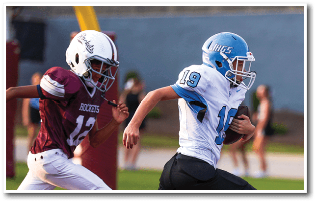 Two people wearing football helmets and uniforms are actively running on a field, with one holding a football and the other in pursuit. Description generated by AI