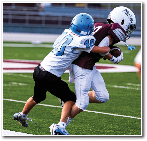 Two people wearing football helmets and uniforms are engaged in a tackle on a football field. Description generated by AI