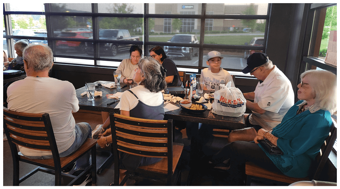 A group of people sitting around a table in a casual dining setting with food and drinks, engaged in conversation and eating. Description generated by AI
