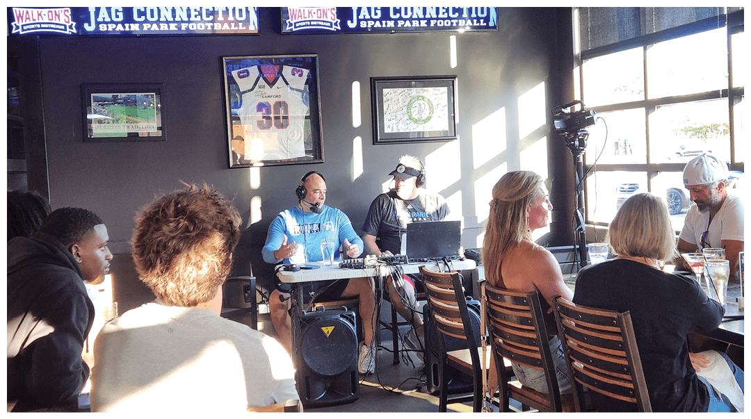 Adults sit at tables and listen to two people speaking into microphones in a casual indoor setting with sports memorabilia on the walls. Description generated by AI