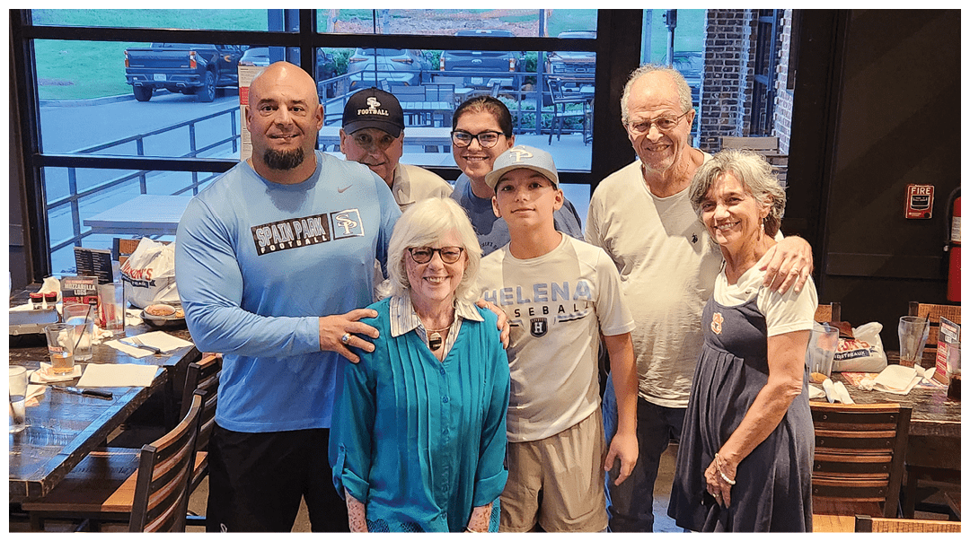 A group of seven people of various ages stand closely together inside a restaurant, smiling and posing for the photo with tables and chairs visible around them. Description generated by AI