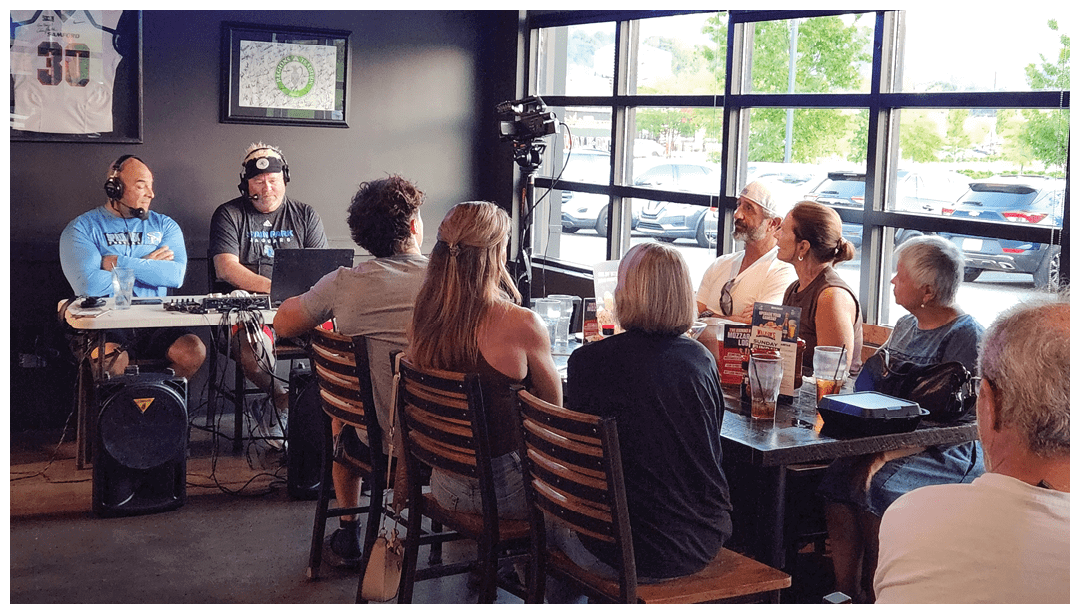 A group of people seated around a table in a casual restaurant setting listen to two adults wearing headphones and speaking into microphones as part of a live broadcast or podcast. Description generated by AI