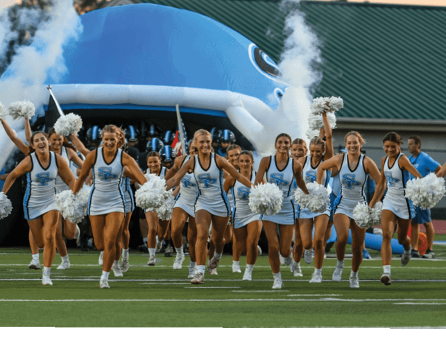 A group of adults in matching cheerleading uniforms with pom-poms are running forward on a sports field with a large inflatable blue whale and smoke in the background. Description generated by AI