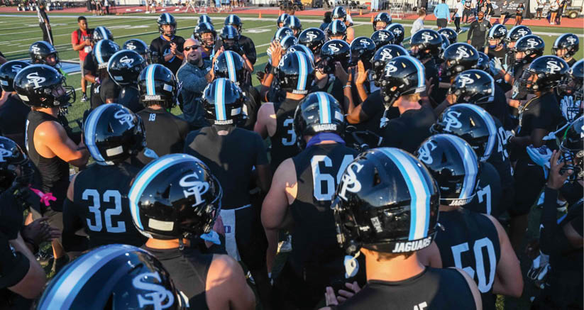 A group of people wearing black football uniforms and helmets with blue and white stripes are gathered closely on a field. Description generated by AI