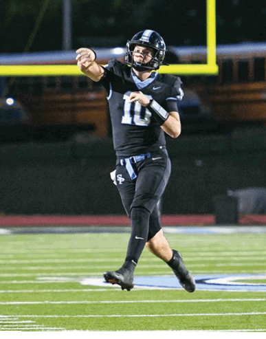 A person wearing a black football uniform is throwing a football on a field with a goalpost and a school bus in the background. Description generated by AI