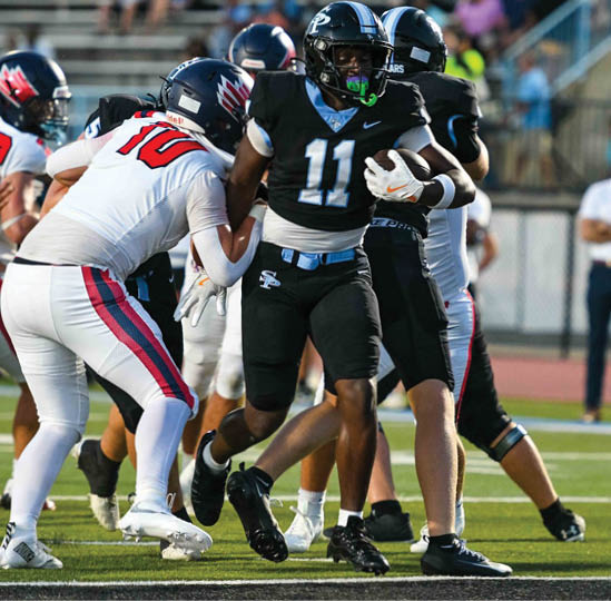 An adult wearing a black football uniform with the number 11 is running on a field while holding a football, with other players and a coach visible in the background. Description generated by AI