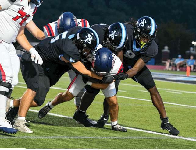 Several people in football uniforms are engaged in a tackle on a grassy field during a game. Description generated by AI