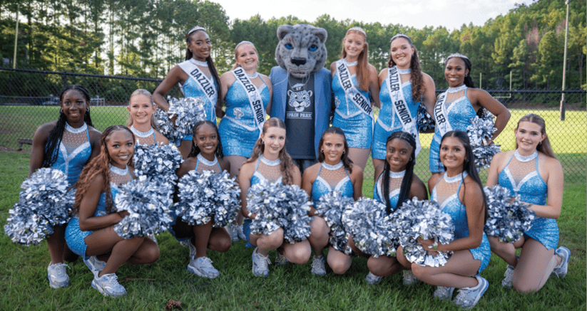 A group of people wearing blue sparkly cheerleading outfits and sashes pose smiling around a person in a gray wildcat mascot costume outdoors with trees in the background. Description generated by AI