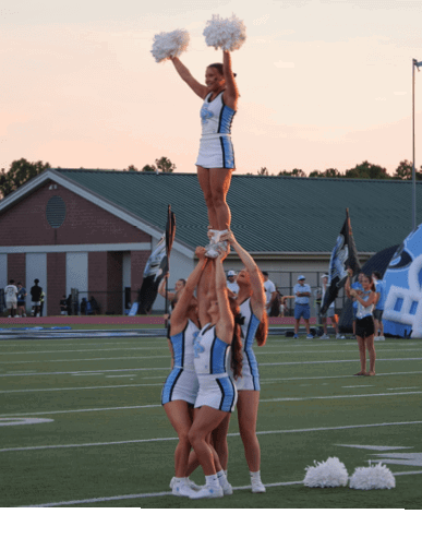 Four adults in cheerleading uniforms perform a stunt on a sports field at sunset with one person lifted high holding pom-poms while others support from below. Description generated by AI