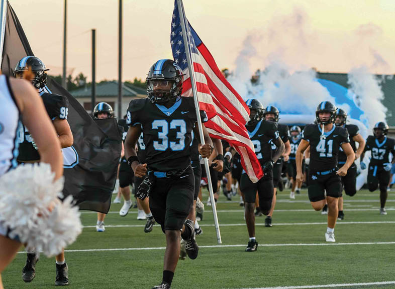 People in black football uniforms are running on a field with one person holding an American flag and smoke rising in the background. Description generated by AI