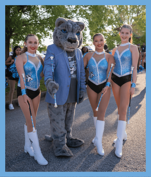 A person wearing a large animal mascot head and a blue suit stands next to a smiling person in a sparkly blue and white dance costume outdoors with trees in the background. Description generated by AI