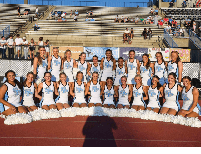 A group of people in matching cheerleading uniforms pose together on a track in front of bleachers with some spectators. Description generated by AI