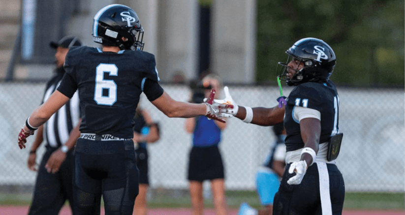 Two people in black football uniforms are giving each other a hand gesture on a sports field with a blurred background. Description generated by AI