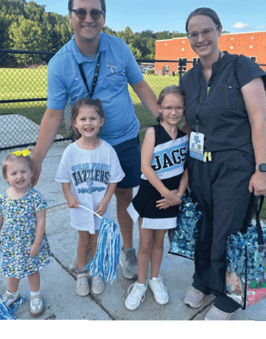 Two adults and two children wearing sports-themed clothing smile outdoors near a fence with trees and a building in the background. Description generated by AI