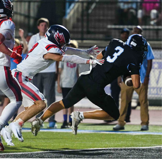 Two people in football uniforms are actively engaged in a play on a field, with one person in black running with the ball while the other in white reaches out to tackle. Description generated by AI
