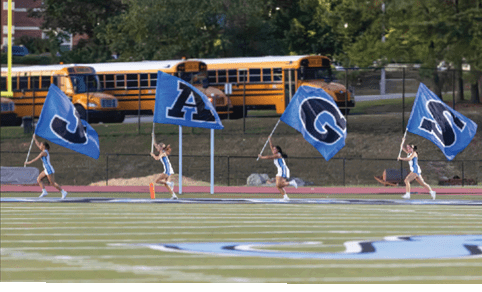Four people running on a sports field each holding a large blue flag with a letter, spelling out the word “JAGS," with school buses and trees in the background. Description generated by AI