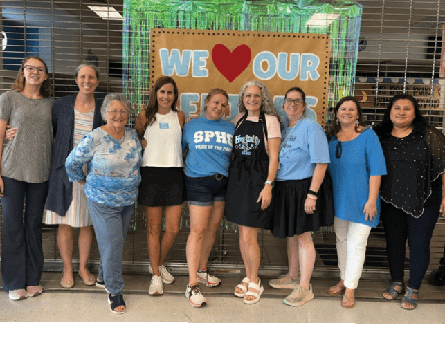 A group of people standing side by side smiling in front of a sign that reads “We love our teachers" with a heart symbol. Description generated by AI
