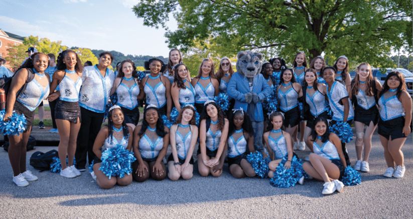 A large group of people dressed in matching blue and black cheerleading outfits pose outdoors on a sunny day with a person in a blue mascot costume standing in the middle. Description generated by AI