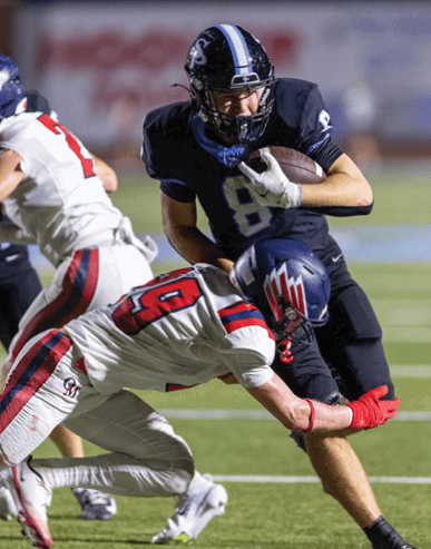 A person in a dark uniform is running with a football while two people in white uniforms with red and blue stripes attempt to tackle them on a football field. Description generated by AI