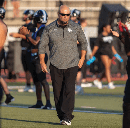 A person wearing sunglasses and a gray long-sleeve shirt walks on a sports field with several people in athletic gear in the background. Description generated by AI