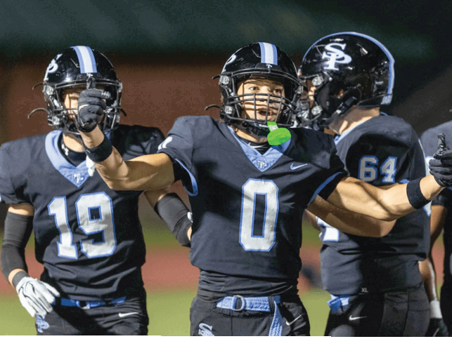 Four people in football uniforms and helmets are on a field with one person in the center raising their arms in celebration or excitement. Description generated by AI