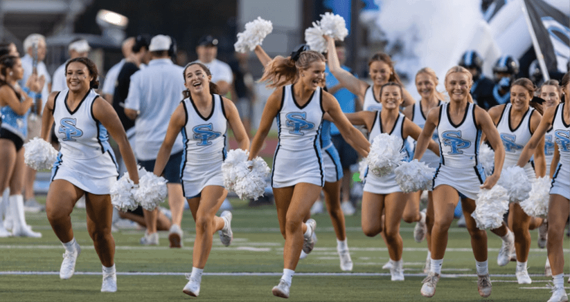 A group of adults in matching cheerleading uniforms are running on a sports field holding white pom-poms with smiles and energetic expressions. Description generated by AI
