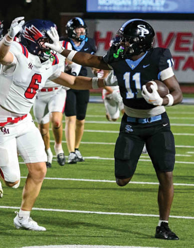 A person in a black football uniform is running with the ball while another person in a white football uniform reaches out to block them on a lit football field. Description generated by AI