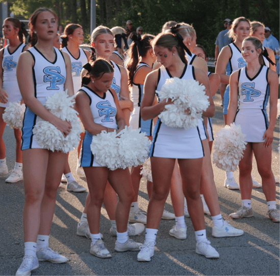 A group of people wearing matching cheerleading uniforms with pom-poms stand outdoors on a paved surface in sunlight. Description generated by AI