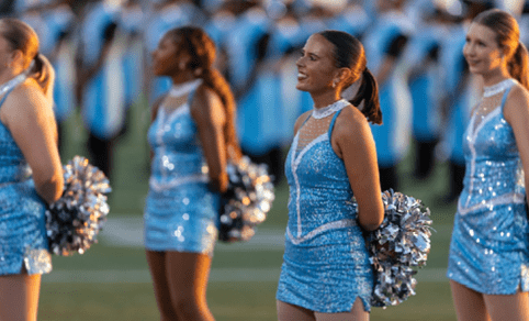 Adults in sparkly blue dance costumes hold silver pom-poms while standing on a field with a blurred marching band in the background. Description generated by AI