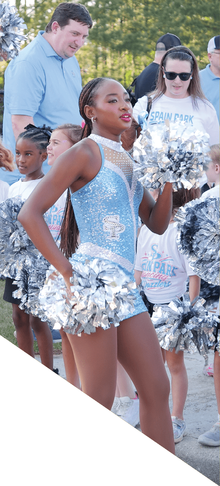 A group of people in sparkly blue cheerleading outfits and others in casual clothing are gathered outdoors on a street and grassy area, holding silver pom-poms. Description generated by AI