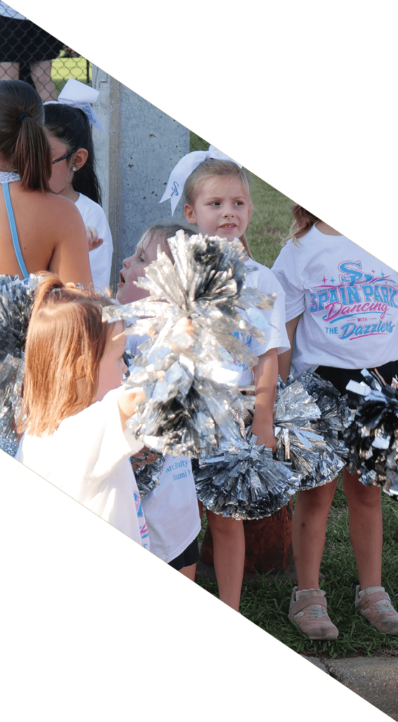Children wearing matching cheerleading outfits hold silver pom-poms while standing outdoors near a fence. Description generated by AI